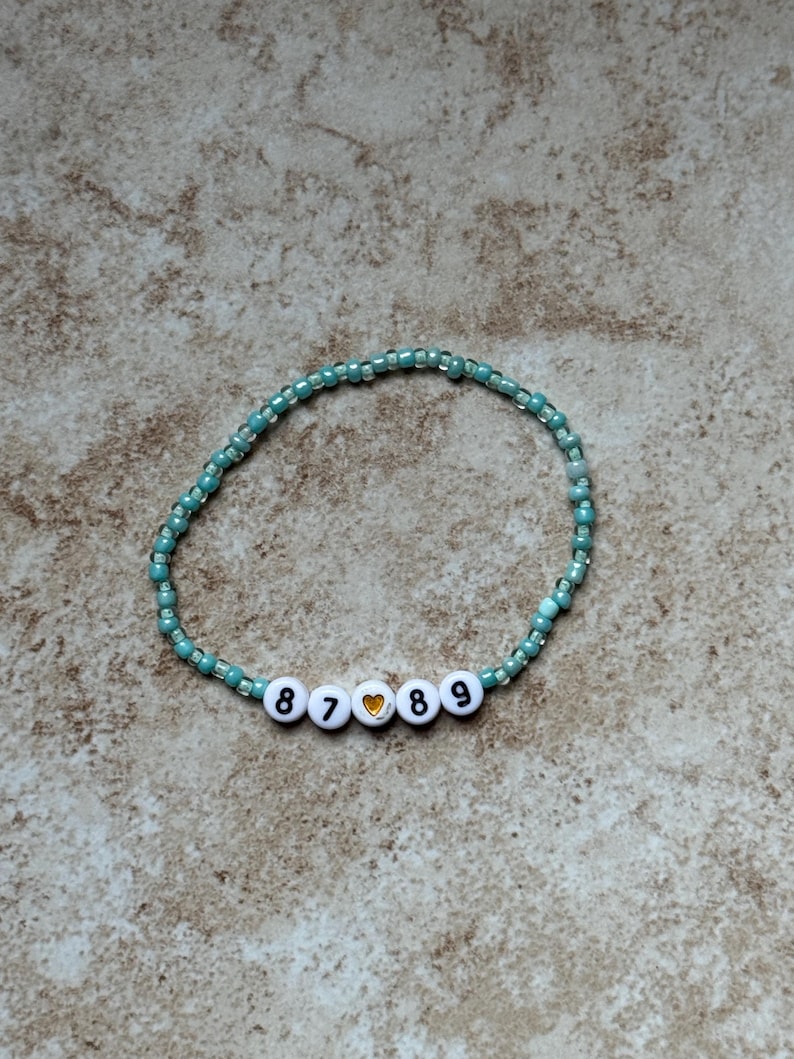 Pode incluir: Uma pulseira de contas turquesa e transparentes. A pulseira apresenta contas de letras brancas que soletram os n&uacute;meros "8789" com uma conta de cora&ccedil;&atilde;o dourado. A pulseira &eacute; exibida em uma superf&iacute;cie de tom neutro.