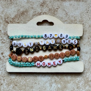Pode incluir: Uma cole&ccedil;&atilde;o de pulseiras de contas com v&aacute;rias frases e n&uacute;meros. As pulseiras apresentam contas de letras brancas, pretas e douradas que soletram frases como "JUST SAY YES" e "END GAME". Algumas pulseiras t&ecirc;m contas turquesas.