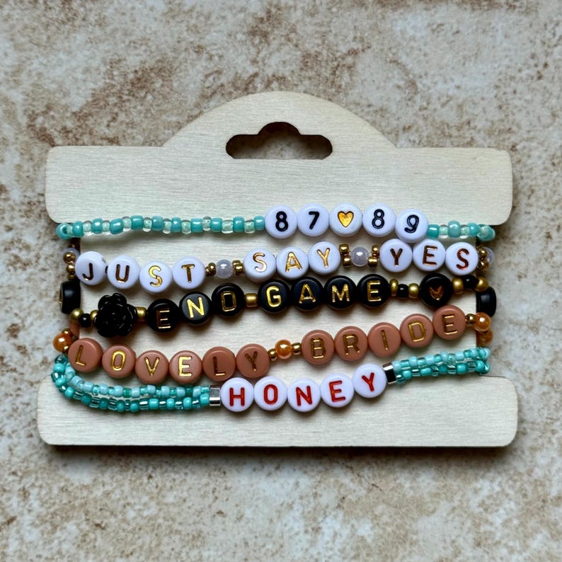 Pode incluir: Uma cole&ccedil;&atilde;o de pulseiras de contas com v&aacute;rias frases e n&uacute;meros. As pulseiras apresentam contas de letras brancas, pretas e douradas que soletram frases como "JUST SAY YES" e "END GAME". Algumas pulseiras t&ecirc;m contas turquesas.