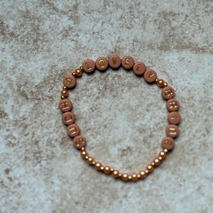 Pode incluir: Uma pulseira com a palavra "LOVELY" escrita em letras douradas em contas cor-de-rosa. A pulseira &eacute; enfiada num cord&atilde;o el&aacute;stico dourado e apresenta contas douradas mais pequenas entre as letras.