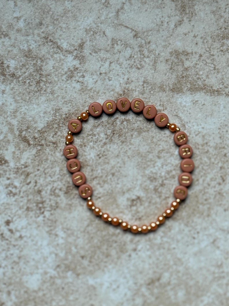Pode incluir: Uma pulseira com a palavra "LOVELY" escrita em letras douradas em contas cor-de-rosa. A pulseira &eacute; enfiada num cord&atilde;o el&aacute;stico dourado e apresenta contas douradas mais pequenas entre as letras.