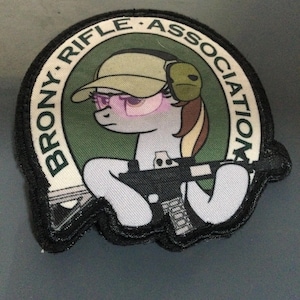 Op de afbeelding: Geborduurde patch met een cartoonpony met een pet, oorbeschermers en een roze bril, die een geweer vasthoudt. De patch is cirkelvormig met de tekst "BRONY RIFLE ASSOCIATION" in witte letters op een groene achtergrond.