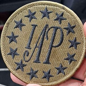May include: A tan circular patch with black embroidered stars around the edge and the letters "IAP" in the center.