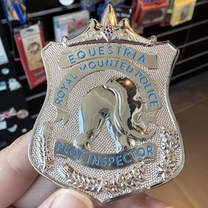 May include: A silver-colored badge with the text "Equestria Royal Mounted Police" and "Plot Inspector" in blue lettering. The badge features a horse silhouette and decorative laurel leaves.
