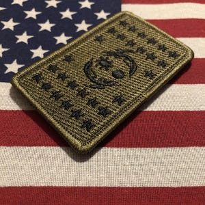 May include: An olive green rectangular patch with a black embroidered design featuring stars and a central emblem. The patch is set against the backdrop of an American flag, with its red and white stripes and blue field of stars.