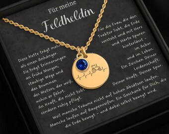 May include: Gold-colored necklace with a round pendant featuring a tractor illustration and a blue gemstone. The necklace is presented in a black box with German text.