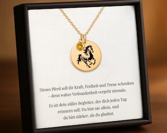 May include: Gold-colored pendant necklace featuring a circular charm with a black horse silhouette. A small yellow gemstone hangs below the chain. The necklace is presented in a black box with text in German.