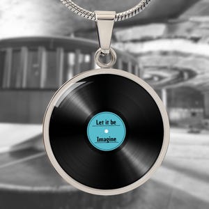 Könnte beinhalten: Eine silberne Halskette mit einem runden Anhänger, der eine schwarze Schallplatte mit dem Text "Let it be" und "Imagine" in einem blauen Kreis zeigt.