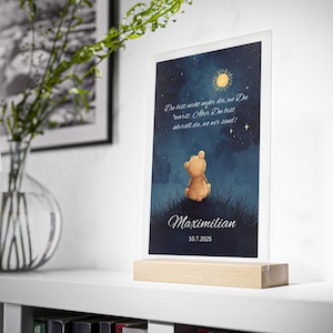 May include: A framed print featuring a watercolor illustration of a teddy bear sitting in grass under a starry night sky. The print includes text in German, the name "Maximilian", and the date 10.7.2025. The frame is clear, and the base is made of wood.