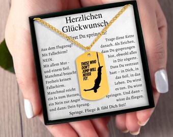 May include: A gold-colored dog tag necklace with a chain. The tag has the text "THOSE WHO DON'T JUMP WILL NEVER FLY" and a silhouette of a person bungee jumping. The tag is presented in a black box with German text.