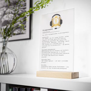 May include: A clear acrylic sign with the word "Disponent" and text in German, set in a wooden base. The sign features a gold and black graphic of a headset with a crown and the number 1.