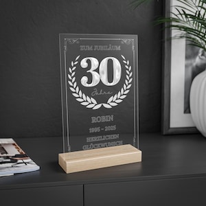 May include: A clear acrylic plaque with the number "30" inside a laurel wreath, mounted on a light wood base. The plaque has text that reads "ZUM JUBILÄUM", "Jahre", "ROBIN 1995-2025", and "HERZLICHEN GLÜCKWUNSCH".