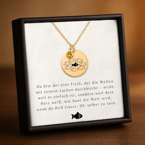 Könnte beinhalten: Eine goldfarbene Halskette mit einem runden Anhänger, der einen Fischschwarm zeigt, von dem einer schwarz ist. Ein kleiner gelber Edelstein befindet sich über dem Anhänger. Die Halskette wird in einer schwarzen Box mit Text präsentiert.