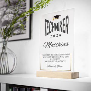 Könnte beinhalten: Transparente Acryl-Abschlussplakette mit dem Wort "TECHNIKER" und dem Jahr "2026" in Schwarz. Der Name "Matthias" steht darunter. Die Plakette steht auf einem hellen Holzsockel. Text unten: "Mama & Papa".