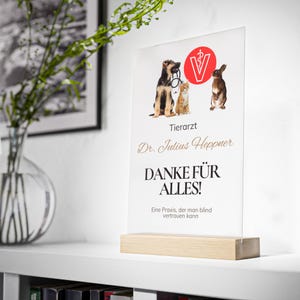 Puede incluir: Un letrero acrílico transparente con una base de madera, con ilustraciones de un perro, un gato y un conejo. El letrero incluye el texto "Tierarzt Dr. Julius Heppner DANKE FÜR ALLES!" y "Eine Praxis, der man blind vertrauen kann."