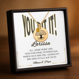 Op de afbeelding: Een goudkleurige ketting met een ronde hanger met een boomontwerp en een kleine gele edelsteen. De ketting wordt gepresenteerd in een zwarte doos met de woorden "YOU GOT IT!" en de naam "Larissa".