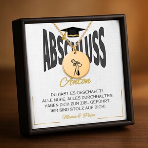 Op de afbeelding: Een gouden afstudeerhalsketting met een ronde hanger met een diploma-illustratie. De ketting wordt gepresenteerd in een zwarte doos met het woord "ABSUSS" en de naam "Anton". De doos bevat ook Duitse tekst.