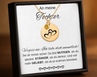 May include: A gold-colored necklace with a round pendant featuring two black hearts. The pendant has a small yellow gemstone and hangs from a delicate chain. The necklace is displayed in a black box with German text.