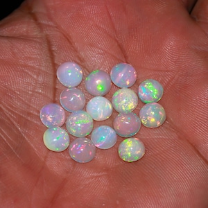 Könnte beinhalten: Eine Ansammlung von dreizehn kleinen, runden, im Cabochon-Schliff geschliffenen Opalen. Jeder Edelstein weist eine durchscheinende, milchig-weiße Basis mit lebhaften, irisierenden Farbblitzen in Grün, Rosa und Blau auf den glatten, gewölbten Oberflächen auf.