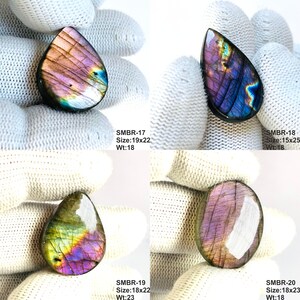 May include: Four polished labradorite gemstones in teardrop and oval shapes. The stones display iridescent colors of purple, blue, and green. Each stone has a unique pattern and is set against a white background. The text "SMBR-17 Size:19x22 Wt:18", "SMBR-18 Size:15x25 Wt:18", "SMBR-19 Size:18x22 Wt:23", and "SMBR-20 Size:18x23 Wt:18" is visible.
