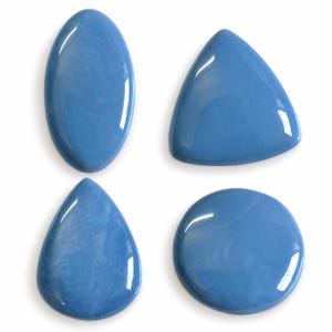 Op de afbeelding: Vier gepolijste, gladde, lichtblauwe cabochons in verschillende vormen, waaronder een ovaal, een driehoek, een druppel en een cirkel. Elk stuk heeft een glanzend, reflecterend oppervlak met subtiele, wolkachtige en gemarmerde blauwe tinten.