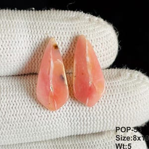 May include: Two teardrop-shaped pink opal gemstones with white veining and small dark inclusions. The stones are labeled POP-53, measuring 8x19 mm, with a weight of 5 carats. The surface of the stones is smooth and polished with a glossy finish.