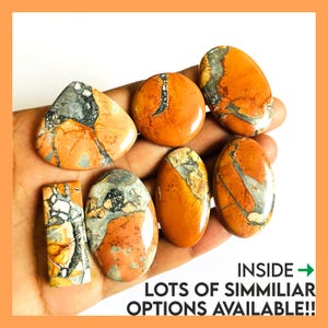 Op de afbeelding: Een verzameling gepolijste edelstenen in tinten oranje, grijs en crème. De stenen hebben verschillende vormen, waaronder ovaal, rond en rechthoekig. De tekst "INSIDE -> LOTS OF SIMMILAR OPTIONS AVAILABLE!!" is zichtbaar.