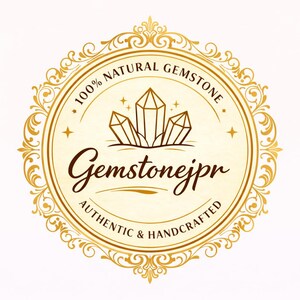 Pu&ograve; includere: Un logo circolare dorato con un bordo in filigrana ornato. Il centro mostra un grafico di tre cristalli con il testo Gemstonejpr, 100% NATURAL GEMSTONE e AUTHENTIC & HANDCRAFTED scritto in un carattere serif e script marrone scuro.