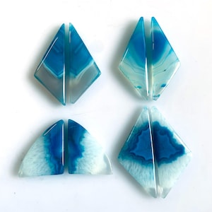 Puede incluir: Ocho rodajas de ágata pulida dispuestas en cuatro pares. Cada pieza presenta un centro azul vibrante con patrones de bandas blancas, translúcidas y grises. Las piedras están cortadas en formas geométricas de diamante y triángulo.