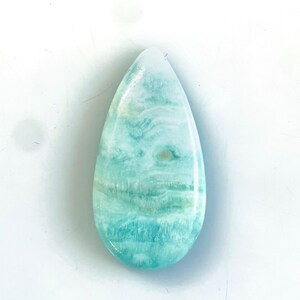 Peut inclure: Une pierre pr&eacute;cieuse Larimar polie en forme de goutte, pr&eacute;sentant une surface lisse et brillante avec des motifs tourbillonnants de bleu clair, turquoise et blanc, rappelant les vagues de l'oc&eacute;an. La pierre a une base arrondie.