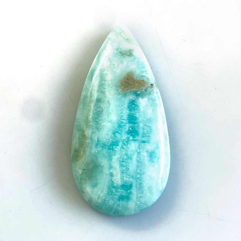 Peut inclure: Une pierre pr&eacute;cieuse polie en forme de goutte d'eau pr&eacute;sentant un motif marbr&eacute; turquoise et blanc avec de subtiles inclusions brun clair. La pierre poss&egrave;de une surface lisse et brillante ainsi qu'une texture vein&eacute;e verticale.