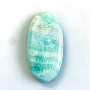 Peut inclure: Une pierre pr&eacute;cieuse polie de forme ovale pr&eacute;sentant une surface lisse avec un motif marbr&eacute; de stries turquoise clair, vert mousse et blanc. La pierre a une finition brillante et affiche un aspect naturel et stratifi&eacute; sur toute sa structure.