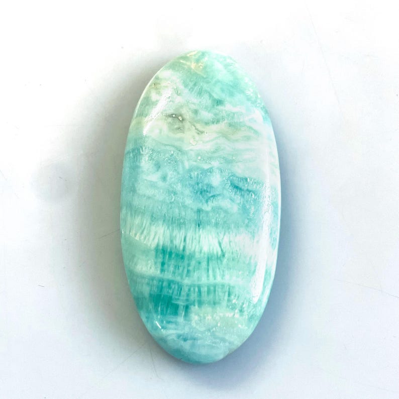 Peut inclure: Une pierre pr&eacute;cieuse polie de forme ovale pr&eacute;sentant une surface lisse avec un motif marbr&eacute; de stries turquoise clair, vert mousse et blanc. La pierre a une finition brillante et affiche un aspect naturel et stratifi&eacute; sur toute sa structure.