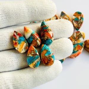 May include: A collection of teardrop-shaped gemstones with a vibrant mix of colors. The stones feature turquoise, orange, and gold hues, with a polished, glossy finish. The gemstones are displayed on a white, textured surface.