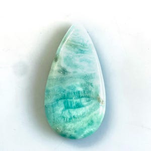 Peut inclure: Une pierre pr&eacute;cieuse polie en forme de goutte d'eau pr&eacute;sentant un motif marbr&eacute; dans des tons bleu clair, turquoise et blanc. La pierre poss&egrave;de une surface lisse et brillante avec des stries horizontales et des variations naturelles.