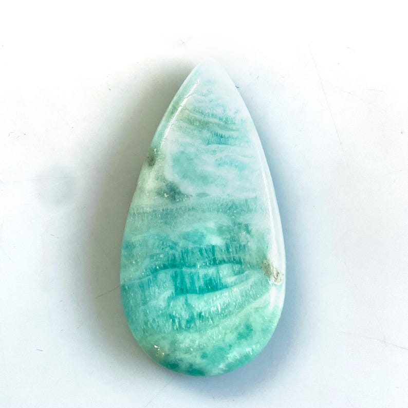 Peut inclure: Une pierre pr&eacute;cieuse polie en forme de goutte d'eau pr&eacute;sentant un motif marbr&eacute; dans des tons bleu clair, turquoise et blanc. La pierre poss&egrave;de une surface lisse et brillante avec des stries horizontales et des variations naturelles.