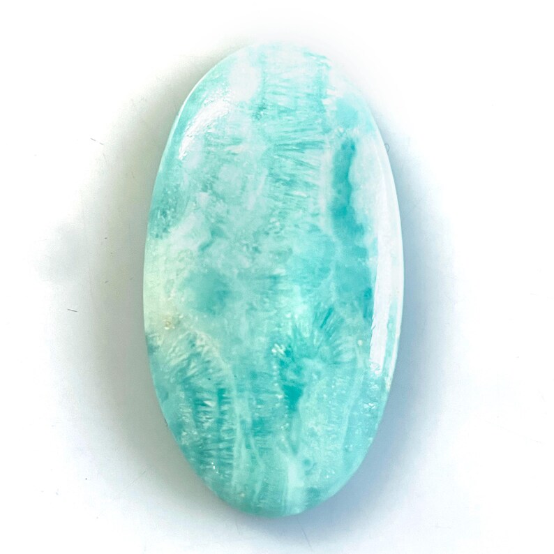 Peut inclure: Une pierre pr&eacute;cieuse cabochon de forme ovale avec une surface polie pr&eacute;sentant un motif marbr&eacute; de stries bleu clair, turquoise et blanches. La pierre affiche une finition lisse et r&eacute;fl&eacute;chissante avec des inclusions nuageuses.