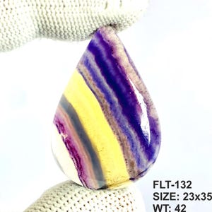 May include: A polished, pear-shaped fluorite gemstone featuring distinct bands of deep purple, bright yellow, and translucent white. The stone is labelled with the text FLT-132, SIZE: 23x35, WT: 42. The surface is smooth and reflective with a glossy finish.
