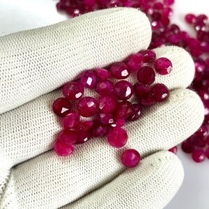 Op de afbeelding: Een verzameling gefacetteerde, ronde, dieprode edelstenen. De edelstenen zijn verspreid over een witte, getextureerde ondergrond. De edelstenen hebben een gepolijst, reflecterend oppervlak en zijn waarschijnlijk robijnen of vergelijkbare edelstenen.