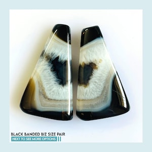 Puede incluir: Un par de piedras de ágata pulidas, triangulares, con bandas blancas y negras, bordes exteriores oscuros y centros blancos cristalinos. El texto en la parte inferior dice: BLACK BANDED BIZ SIZE PAIR. NEXT TO SEE MORE OPTIONS.