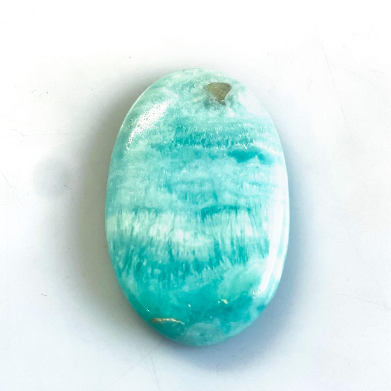 Peut inclure: Une pierre pr&eacute;cieuse polie de forme ovale pr&eacute;sentant une palette de couleurs turquoise et sarcelle vibrante avec des inclusions blanches distinctes en forme de nuages et une texture de surface lisse et brillante qui refl&egrave;te la lumi&egrave;re.