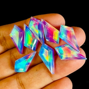 Op de afbeelding: Een verzameling iriserende, vlieger-vormige edelstenen. De edelstenen vertonen een spectrum aan kleuren, waaronder blauw, groen, roze en geel, met een gepolijste, gefacetteerde uitstraling. De edelstenen worden in een hand tegen een donkere achtergrond gehouden.