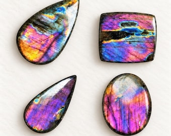 Peut inclure: Quatre pierres précieuses de labradorite polies en forme de goutte, de rectangle et d'ovale. Chaque pierre présente des reflets irisés aux couleurs vives violettes, bleues et jaune doré sur des surfaces lisses et réfléchissantes avec des inclusions minérales.