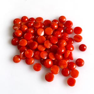 May include: A collection of numerous small, round, flat-backed cabochons in a vibrant, solid orange-red color. Each piece features a smooth, polished surface with a glossy finish, arranged in a loose, scattered pile against a plain white background.