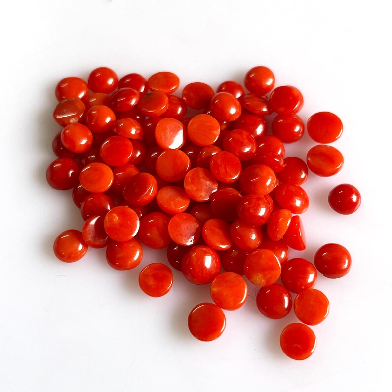May include: A collection of numerous small, round, flat-backed cabochons in a vibrant, solid orange-red color. Each piece features a smooth, polished surface with a glossy finish, arranged in a loose, scattered pile against a plain white background.