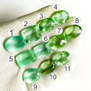 Puede incluir: Once piedras preciosas verdes facetadas dispuestas sobre una superficie texturizada, cada una marcada con un número blanco en un círculo del uno al once. La colección incluye una mezcla de cortes ovalados y rectangulares en tonos verdes.