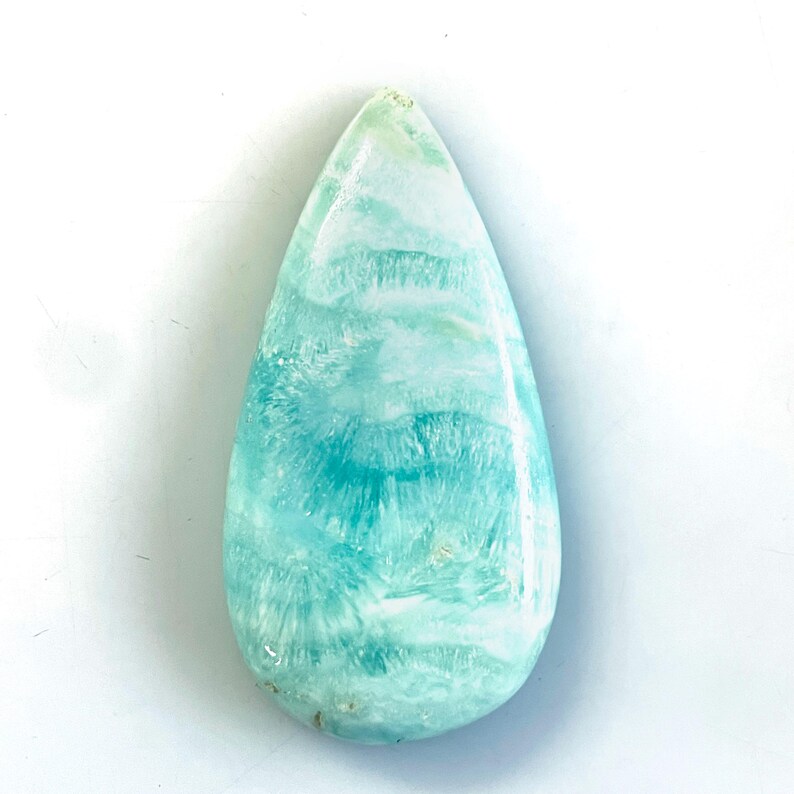 Peut inclure: Une pierre pr&eacute;cieuse polie en forme de goutte d'eau pr&eacute;sentant un m&eacute;lange vibrant de turquoise, de sarcelle et de blanc. La pierre affiche un motif fibreux rayonnant distinct sur toute sa surface, avec une finition lisse et brillante.