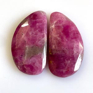 May include: Two faceted, teardrop-shaped gemstones in shades of deep pink and magenta. The stones have a polished, reflective surface with visible internal patterns and inclusions. The gemstones are set against a white background.