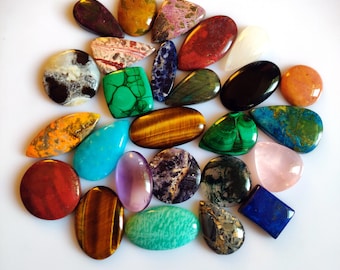 Può includere: Una collezione di pietre preziose lucide di varie forme e colori, tra cui ovali, a goccia e irregolari. Le pietre preziose presentano una gamma di tonalità, come malachite verde, occhio di tigre, ametista e quarzo rosa. La disposizione è su una superficie bianca.