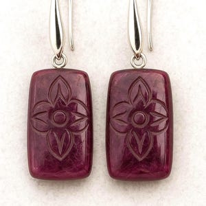 Peut inclure: Une paire de boucles d'oreilles pendantes rectangulaires avec des pierres magenta foncé gravées d'un motif floral à quatre pétales. Les pierres sont suspendues à des crochets en métal argenté poli, offrant un design net et symétrique.